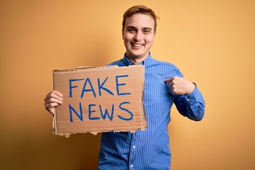 Young handsome redhead man holding banner with fake news message over yellow background pointing...