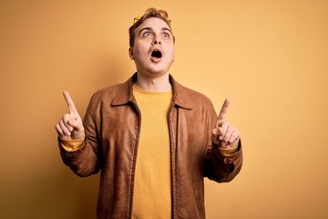 Young handsome redhead man wearing casual leather jacket over isolated yellow background amazed and surprised looking up and pointing with fingers and raised arms.