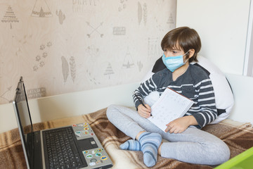 Distance learning for children during the coronavirus epidemic. The boy sits on the bed and receives a school assignment using the Internet and a laptop.