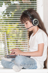 A child is working on a laptop by the window. A little girl (seven years old) is sitting on a windowsill by the window and working on a laptop. One person