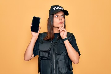 Beautiful brunette policewoman wearing police uniform holding smartphone showing screen serious face thinking about question, very confused idea