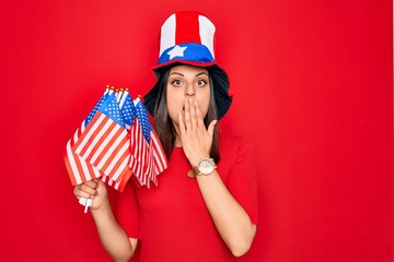 Young beautiful brunette patriotic woman wearing united states hat holding usa flags covering mouth...