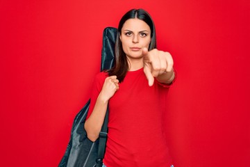 Young beautiful brunette guitarist woman holding guitar case over isolated red background pointing with finger to the camera and to you, confident gesture looking serious