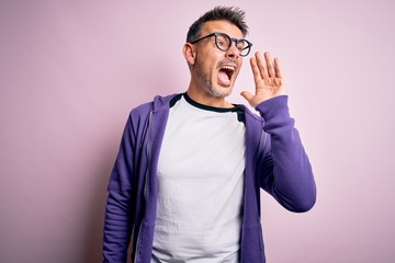 Young handsome man wearing purple sweatshirt and glasses standing over pink background shouting and screaming loud to side with hand on mouth. Communication concept.