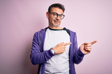 Young handsome man wearing purple sweatshirt and glasses standing over pink background Pointing aside worried and nervous with both hands, concerned and surprised expression