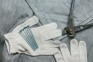 A handful of metallic self-cutters screws and very aged and rusty pliers lies on Safety knitted Work Gloves for industrial use against the backdrop of an old denim fabric. Selective focus macro shot