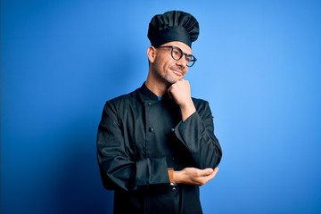 Young handsome chef man wearing cooker uniform and hat over isolated blue background with hand on chin thinking about question, pensive expression. Smiling and thoughtful face. Doubt concept.
