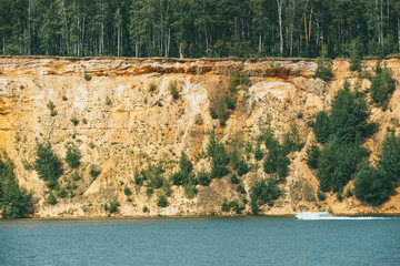 High rocky sandy steep bank covered with pine trees over a clear and blue lake. fast motor boat cuts through the water surface