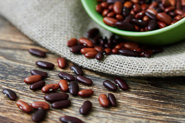 A mix of the red beans and roman beans sprinkled on the rustic wooden table from a green bowl standing on the canvas. Horizontal photo. Copy space, space for text