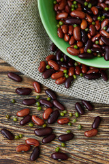 A mix of red beans, mung beans (mash) and roman beans sprinkled on the rustic wooden table from a green bowl standing on the canvas. Top view. Vertical photo