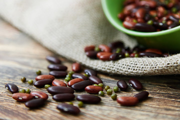 A mix of red beans, mung beans (mash) and roman beans sprinkled on the rustic wooden table from a green bowl standing on the canvas. Copy space, space for text. Horizontal photo