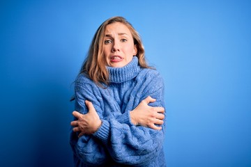Young beautiful blonde woman wearing casual turtleneck sweater over blue background shaking and freezing for winter cold with sad and shock expression on face