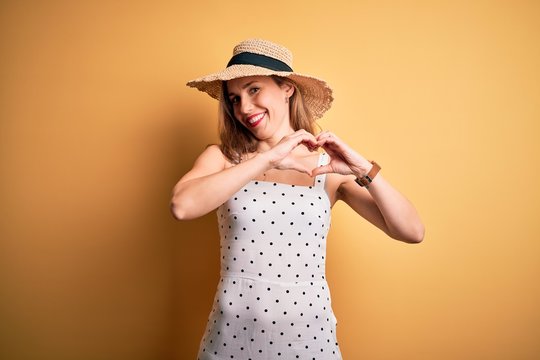 Young beautiful blonde woman on vacation wearing summer hat over yellow background smiling in love showing heart symbol and shape with hands. Romantic concept.