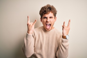 Young blond handsome man with curly hair wearing casual sweater over white background shouting with crazy expression doing rock symbol with hands up. Music star. Heavy concept.