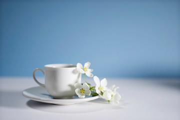 Fototapeta na wymiar White cup of morning espresso with a blossoming apple tree branch on a stone surface of a table with reflections opposite blue background.