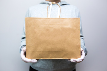 A delivery service worker in gloves, on a gray background, is holding a paper craft bag from the store. Delivery service concept. Infection protection COVID-19