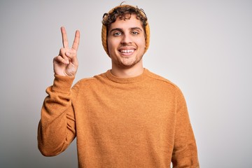 Young blond handsome man with curly hair wearing casual sweater and wool hat smiling with happy face winking at the camera doing victory sign. Number two.