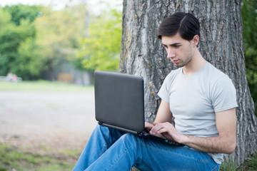 A young caucasian man sitting under a tree in a park and typing on a laptop. Remote work