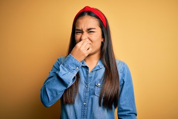 Young beautiful asian woman wearing casual denim shirt and diadem over yellow background smelling something stinky and disgusting, intolerable smell, holding breath with fingers on nose. Bad smell