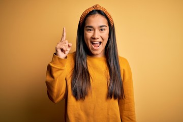 Young beautiful asian woman wearing casual sweater and diadem over yellow background pointing finger up with successful idea. Exited and happy. Number one.