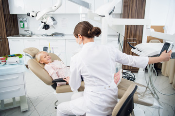 Obraz premium Skillful dentist is examining the teeth of a child. The woman is sitting near the girl trying to set up a contact with her to make her feel comfortable