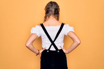 Young beautiful blonde german woman with blue eyes wearing traditional octoberfest dress standing backwards looking away with arms on body