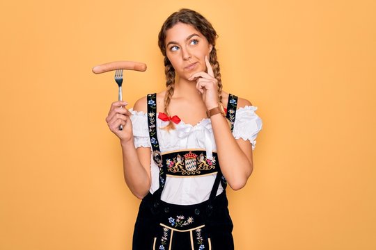 Beautiful german woman with blue eyes wearing oktoberfest dress holding fork with sausage serious face thinking about question, very confused idea