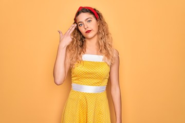 Beautiful blonde pin-up woman with blue eyes wearing diadem standing over yellow background Shooting and killing oneself pointing hand and fingers to head like gun, suicide gesture.