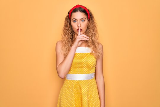 Beautiful blonde pin-up woman with blue eyes wearing diadem standing over yellow background asking to be quiet with finger on lips. Silence and secret concept.