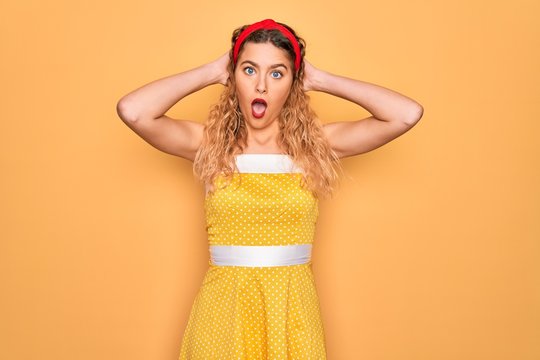 Beautiful blonde pin-up woman with blue eyes wearing diadem standing over yellow background Crazy and scared with hands on head, afraid and surprised of shock with open mouth