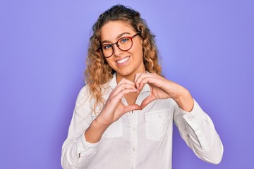 Young beautiful woman with blue eyes wearing casual shirt and glasses over purple background smiling in love doing heart symbol shape with hands. Romantic concept.