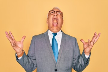 Middle age senior grey-haired handsome business man wearing glasses over yellow background crazy and mad shouting and yelling with aggressive expression and arms raised. Frustration concept.