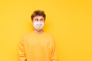 Funny young man in a protective gauze mask stands on a yellow background and looks thoughtfully to the side on copy space. The blond in a medical mask is protected from the coronavirus.