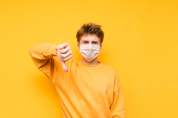 Portrait of a displeased young man in a gauze medical mask stands on a yellow background and shows a finger down. Guy is tired of quarantine. Coronavirus pandemic. Quarantine. covid-19.