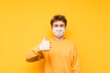 Funny guy in a protective mask stands on a yellow background, looking at the camera with a shocked face and showing a thumbs up. Coronavirus pandemic. Quarantine. covid-19.