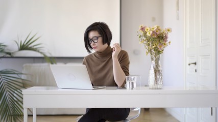 Pan shot right to left of smiling asian woman in eyeglasses working with laptop. Glass of water on table and a vase with flowers in white interior home office