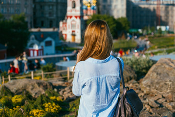 A beautiful red-haired girl in a blue denim shirt stands on a hill in the Park and looks at the opening perspective with people walking and growing bushes. Rear view. Social Distancing concept