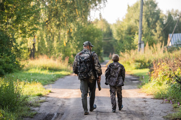 Hunters with hunting equipment going away through rural field towards forest at sunset during hunting season in countryside.