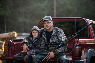 Fototapeta premium Man at his truck with his son in the forest. Hunter teaches young boy how to use shotgun rifle.