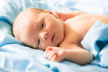 little caucasian newborn baby boy laying down on delicate blue blanket, falling asleep in safe, warm fabric. Adorable, sleepy new born baby  opening his eyes.