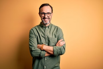 Middle age hoary man wearing casual green shirt and glasses over isolated yellow background happy face smiling with crossed arms looking at the camera. Positive person.