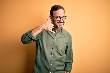 Obraz premium Middle age hoary man wearing casual green shirt and glasses over isolated yellow background smiling doing phone gesture with hand and fingers like talking on the telephone. Communicating concepts.