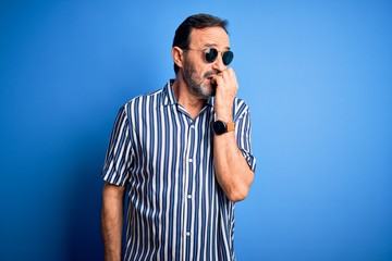 Middle age hoary man wearing striped shirt and sunglasses over isolated blue background looking stressed and nervous with hands on mouth biting nails. Anxiety problem.