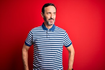 Middle age hoary man wearing casual striped polo standing over isolated red background smiling looking to the side and staring away thinking.
