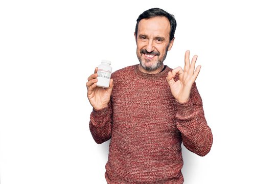 Middle age handsome man holding bottle of medicine pills over isolated white background doing ok sign with fingers, smiling friendly gesturing excellent symbol