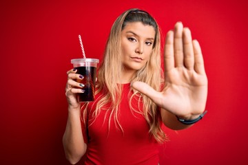 Young beautiful blonde woman drinking cola fizzy beverage using straw over red background with open...