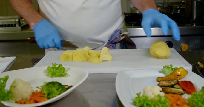 close-up of men's hands in blue disposable gloves. the cook has cut one potato , begins to slice another, and the third is lying on the cutting Board. in the foreground are two plates of vegetables