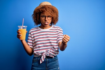 Young African American woman with curly hair on vacation wearing hat drinking orange juice annoyed and frustrated shouting with anger, crazy and yelling with raised hand, anger concept