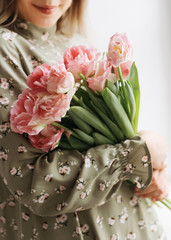 Fototapeta premium young cheerful girl in a dress with a floral pattern with a bouquet of pink Terry tulips in her hands under natural lighting at a large window in a light minimalistic interior