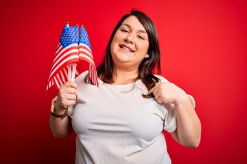 Beautiful plus size woman holding united states of america flag over red background with surprise face pointing finger to himself